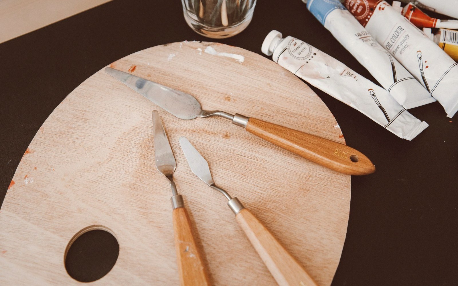 Palette knives resting on a wooden mixing board beside tubes of oil paint