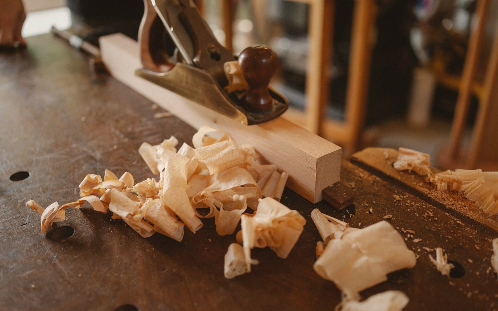 A hand plane rests on a wooden board surrounded by curled wood shavings on a workbench